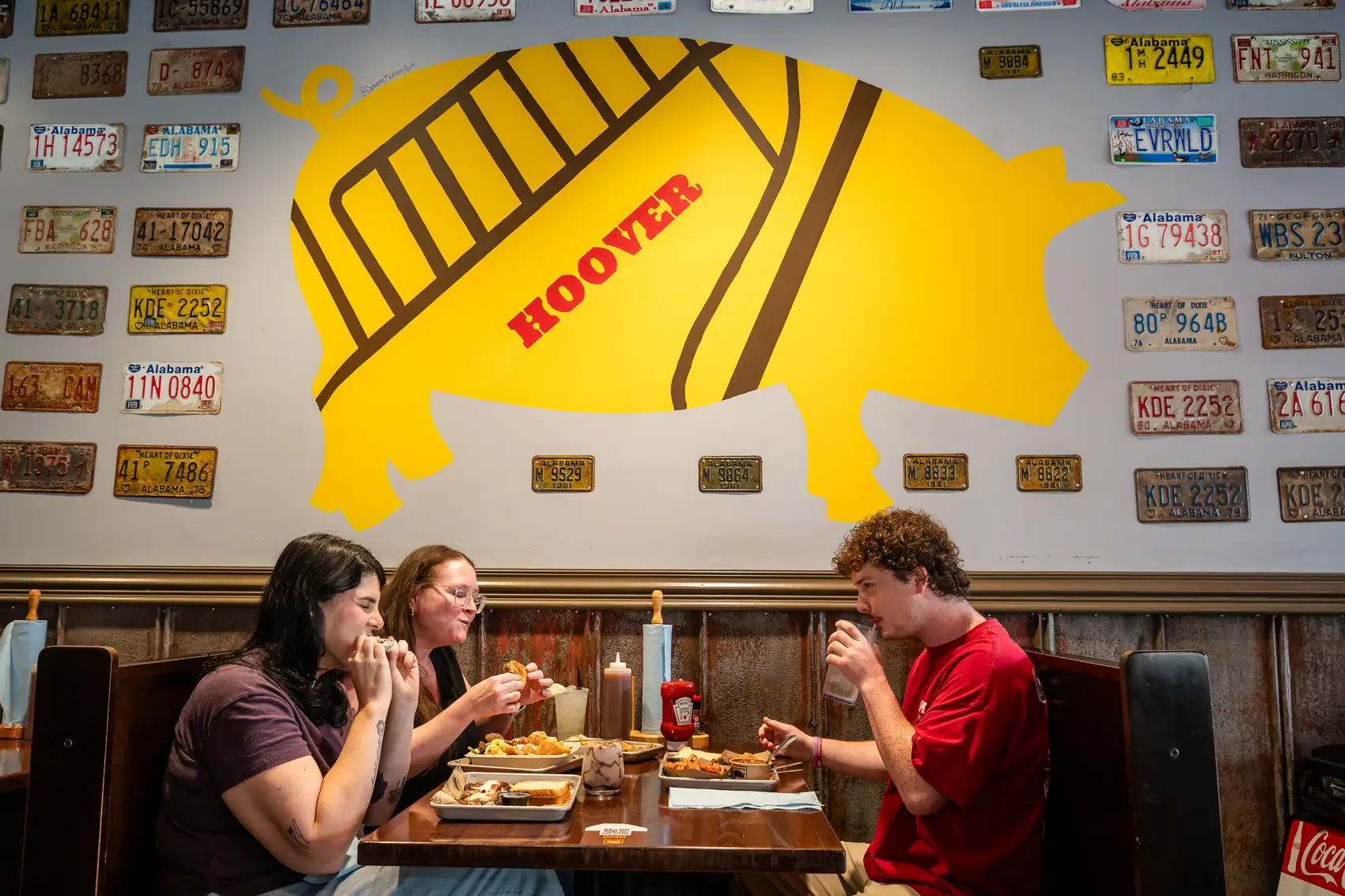 Guests sharing a meal in a SAW's BBQ booth beneath the pig mural and license plate wall